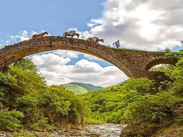 Persona caminando con caballos por un puente de piedra en un valle verde y frondoso.