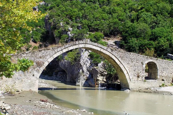 Puente de piedra que cruza un río rodeado de árboles y colinas rocosas.