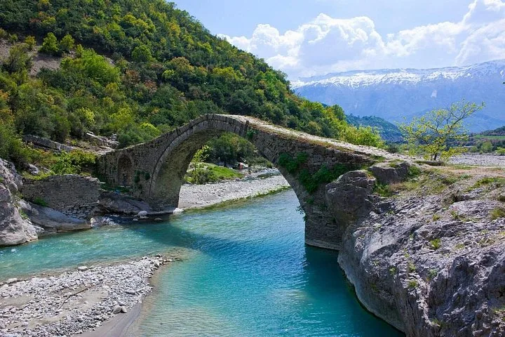 Stone bridge over a blue river with mountains in the background.