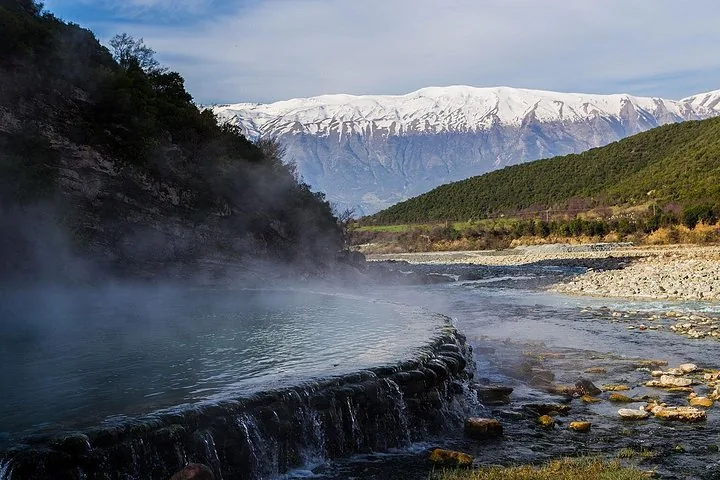 Dampfendes Thermalbecken neben einem Fluss mit schneebedeckten Bergen im Hintergrund.