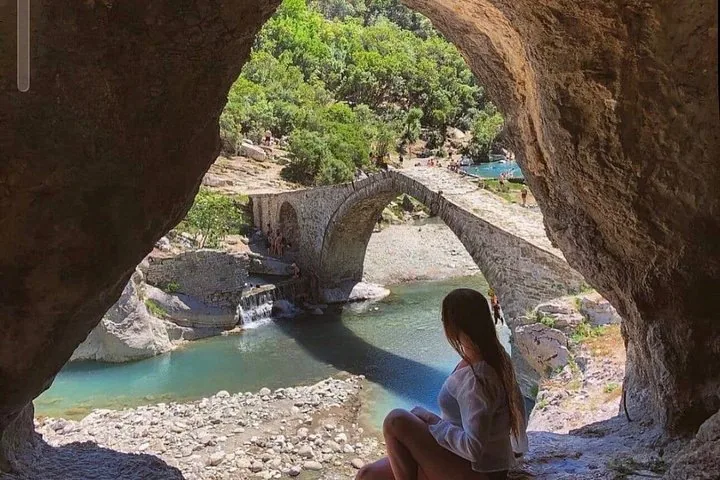 Mujer sentada en una cueva mirando un puente de piedra sobre un río.